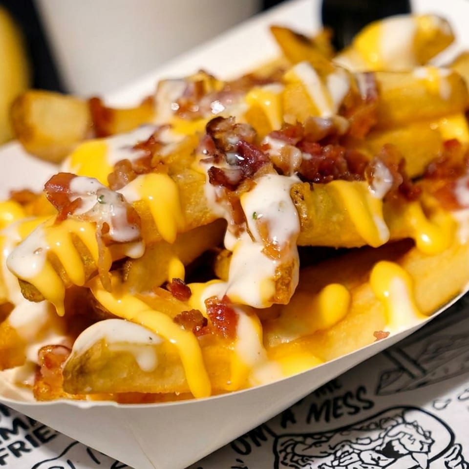 Loaded fries topped with melted cheddar, white sauce, bacon, and chopped toppings in a paper tray.