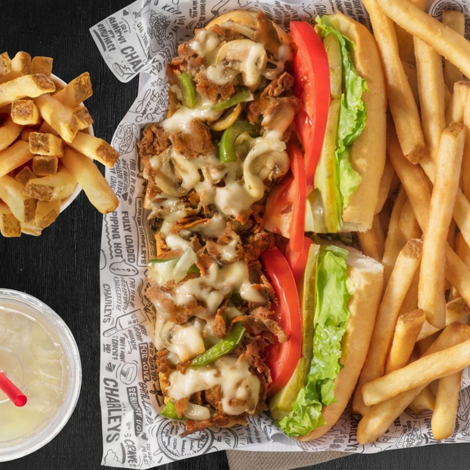 Cheesesteak sandwich with melted cheese, peppers, onions, lettuce, tomato, fries, and a drink on Charley’s branded paper.