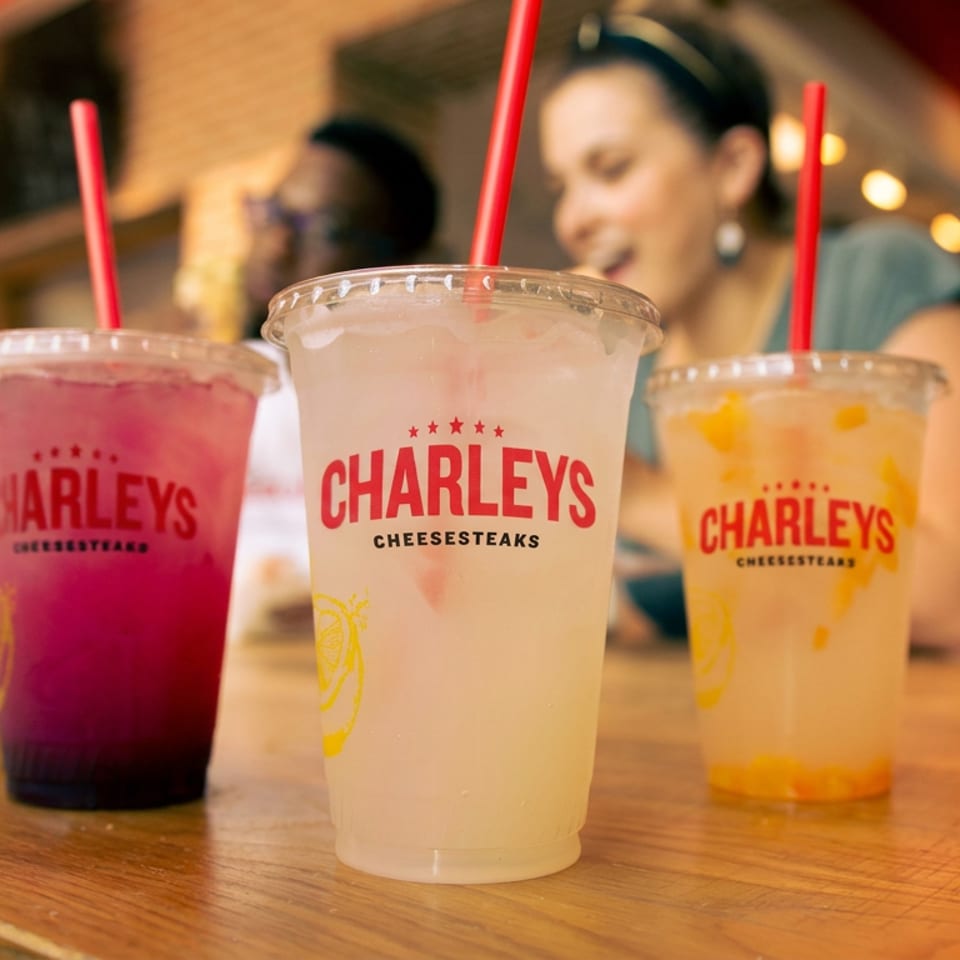Three colorful Charleys Cheesesteaks drinks with red straws sit on a wooden table in a warmly lit restaurant.