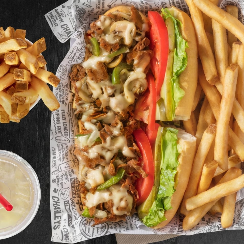 Cheesesteak sandwich with peppers, onions, tomato, and lettuce, surrounded by French fries and a dipping sauce on a dark table.